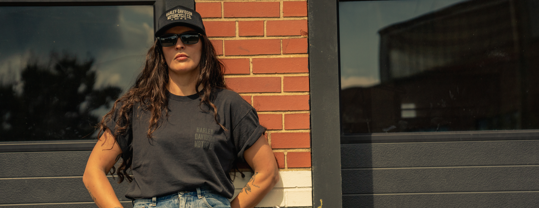 Woman leaning against a building wearing H-D Cap and T-shirt wtih blue jeans and a backpack on strapped on to her harley-davidson motorcycle