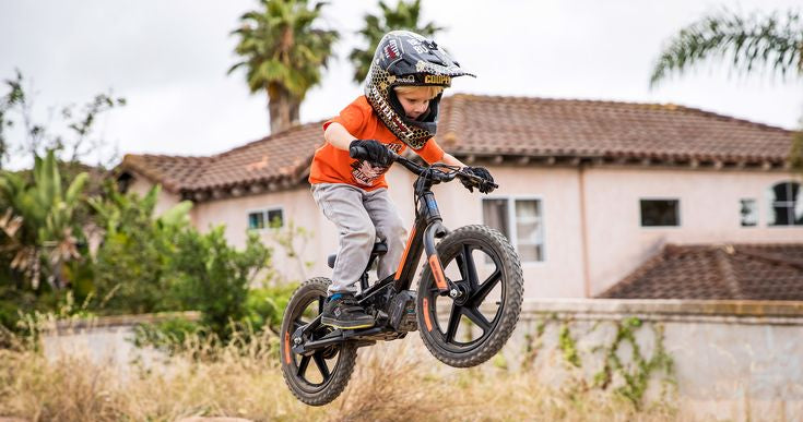 Child on a Harley-davidson balance bike performing a jump in an outdoor setting with houses and trees in the background.